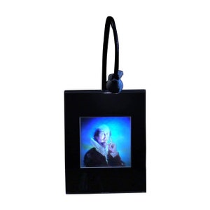 May include: A black framed picture with a portrait of a man in a blue and white patterned shirt. The picture is hanging from a black cord with a black knob.