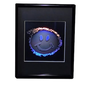 May include: A framed artwork featuring a smiley face design. The smiley face has blue eyes and a curved mouth, outlined in a neon blue glow. The outer edge of the design has a gradient of orange and blue. The frame is black.