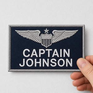 Könnte beinhalten: Ein rechteckiger, dunkelblauer bestickter Aufnäher mit silbernem Rand. Das Design zeigt ein silbernes geflügeltes Schildelement mit einem Stern darüber sowie den weißen gestickten Text CAPTAIN JOHNSON, der unter dem Emblem zentriert ist.
