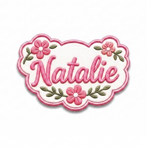 Può includere: Una toppa ricamata bianca con bordo rosa smerlato che presenta il nome Natalie in caratteri corsivi rosa. Il design è decorato con tre fiori rosa con centri bianchi e foglie verdi disposte attorno al testo centrale.