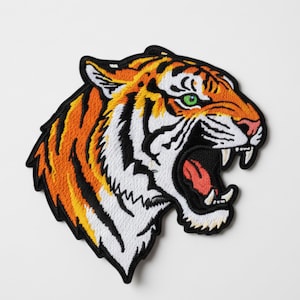 Op de afbeelding: Geborduurde tijgerkop patch met oranje, zwarte en witte strepen en een brullende bek. De patch heeft groene ogen en een zwarte rand, geschikt voor jassen, tassen of andere stoffen items.