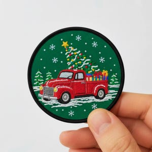 Könnte beinhalten: Gestickter Aufnäher mit einem roten Oldtimer-Truck, der einen Weihnachtsbaum und Geschenke transportiert. Der Aufnäher hat einen grünen Hintergrund mit Schneeflocken und einen schwarzen Rand. Ein festliches Weihnachtsdesign.