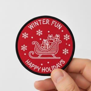Può includere: Una toppa rotonda rossa con bordo nero, con testo e illustrazioni bianche. Il testo recita "Winter Fun" e "Happy Holidays". Una slitta bianca piena di regali è al centro, circondata da fiocchi di neve.