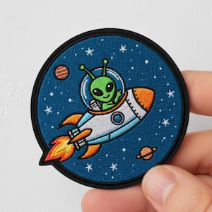 Op de afbeelding: Ronde geborduurde patch met een groene alien in een raket tegen een blauwe sterrenachtergrond. De raket is wit en oranje met een rode en gele vlam. De patch heeft een zwarte rand.