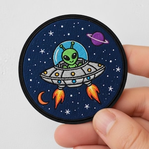 May include: Round embroidered patch featuring a green alien piloting a silver and white UFO against a dark blue starry background. The patch includes a crescent moon, a purple planet, and orange flames.