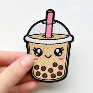 Könnte beinhalten: Gestickter Aufnäher mit einer niedlichen Cartoon-Boba-Tasse mit einem lächelnden Gesicht, schwarzen Augen und rosa Wangen. Die Tasse ist beige mit braunen Boba-Perlen und einem rosa Strohhalm. Der Aufnäher hat einen schwarzen Rand und einen weißen Deckel.