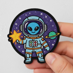 May include: Embroidered patch featuring an alien astronaut in a blue spacesuit, holding a yellow star. The patch has a purple background with stars and a planet with rings. The patch is round with a black border.