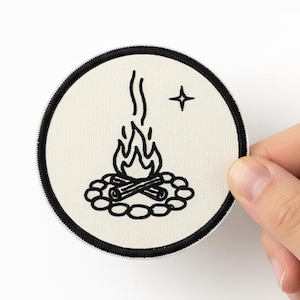 Può includere: Una toppa rotonda color crema con bordo nero. La toppa presenta un'illustrazione nera di un falò con fiamme, tronchi e una stella. La toppa è tenuta da una mano su sfondo bianco.