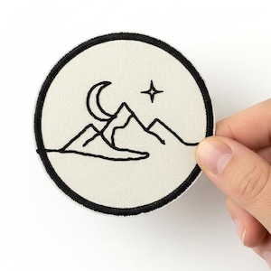 May include: A round patch with a black embroidered design of mountains, a crescent moon, and a star. The patch has a cream-colored background and a black border. The patch is held by a hand.