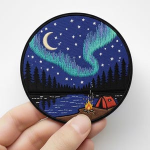 Puede incluir: Parche bordado redondo que representa una escena de acampada nocturna. El diseño incluye un cielo azul con una luna creciente, estrellas y aurora boreal. Una fogata y una tienda roja son visibles cerca de un lago y un bosque.