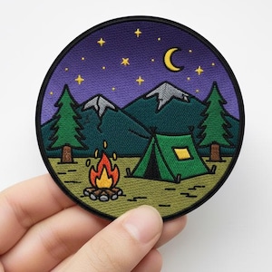 Puede incluir: Un parche bordado circular con una escena de acampada. El diseño incluye una tienda de campaña verde, una fogata, pinos, montañas, una luna creciente y estrellas sobre un cielo nocturno púrpura. El parche está sostenido por una mano.