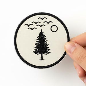 May include: Round patch with a black and white design featuring a pine tree, birds, and a sun. The patch has a black border and is held by a hand against a white background.