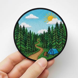 Puede incluir: Parche bordado redondo con un paisaje forestal con un sendero, árboles verdes, una tienda de campaña azul y un sol. El parche tiene un borde negro. Ideal para ropa o bolsos. Perfecto para los amantes del aire libre.