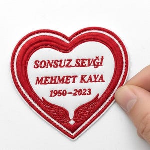 Può includere: Una toppa a forma di cuore con bordo rosso e centro bianco. La toppa presenta il testo "SONSUZ SEVGİ MEHMET KAYA 1950-2023" in rosso, con piccoli disegni di ali in basso. La toppa è tenuta da una mano.