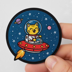 May include: A circular embroidered patch depicting a cartoon cat in a spacesuit, piloting a red spaceship. The cat is smiling against a dark blue starry background with planets. The patch has a black border.