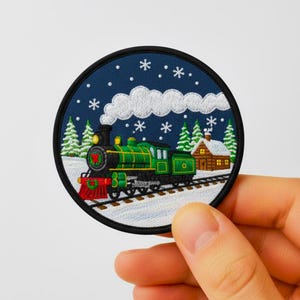 May include: Round embroidered patch depicting a green and black steam train in a snowy scene with a cabin. The patch has a black border, set against a dark blue background with white snowflakes. The design evokes a winter theme.