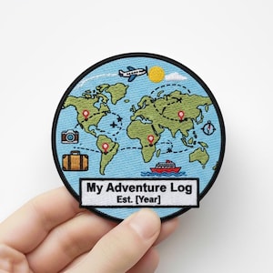 Może przedstawiać: Okrągła naszywka z haftem przedstawiająca mapę świata i ikony podróżnicze. Naszywka zawiera samolot, aparat fotograficzny, walizkę i statek. Tekst na naszywce brzmi "My Adventure Log Est. [Year]" w białym prostokącie.