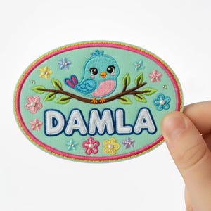Könnte beinhalten: Ovaler, gestickter Aufnäher mit türkisem Hintergrund, der einen blauen Vogel auf einem Ast zeigt. Der Name "DAMLA" ist weiß gestickt. Der Aufnäher ist mit Blumen und einem rosa-grünen Rand verziert.