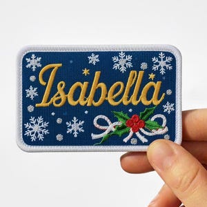Puede incluir: Un parche rectangular azul marino bordado con el nombre "Isabella" en letras doradas. El diseño incluye copos de nieve blancos, una ramita de acebo con bayas rojas y un borde blanco. El parche está sostenido por una mano.