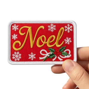 Puede incluir: Un parche rectangular rojo con la palabra "Noel" en letras doradas. El parche presenta copos de nieve blancos y un diseño de acebo verde y rojo. El parche tiene un borde blanco.