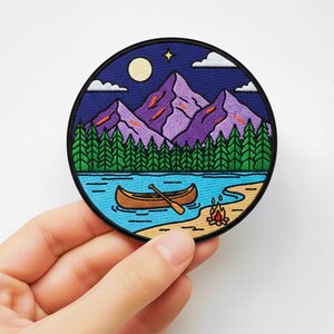 May include: A round embroidered patch featuring a mountain landscape with a lake, canoe, and campfire. The design includes purple mountains, green trees, a blue lake, and a night sky with a moon and stars. The patch has a black border.