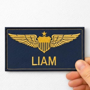 May include: A navy blue patch with gold embroidered wings, a shield, and the name "LIAM". The patch is rectangular and has a gold border. The wings are spread out on either side of the shield.