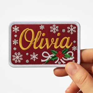 May include: A rectangular, embroidered patch with the name "Olivia" in gold script. The patch is red with white snowflakes and a holly and bow design. The patch is held by a hand.