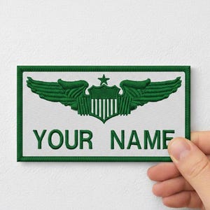 Pode incluir: Patch bordado com um design verde e branco. O patch apresenta um brasão central com uma estrela e asas, e o texto "YOUR NAME" em verde. O patch é retangular com uma borda verde.