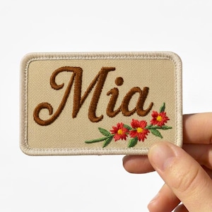 Puede incluir: Parche rectangular beige bordado con el nombre "Mia" en escritura marrón. El parche tiene un borde marrón y presenta flores bordadas rojas y amarillas con hojas verdes.