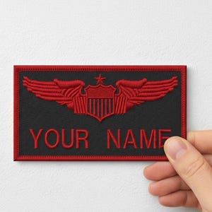 Pode incluir: Patch retangular preto com asas bordadas em vermelho e um desenho de escudo. O texto "YOUR NAME" é bordado em vermelho abaixo do emblema. O patch é segurado por uma mão.