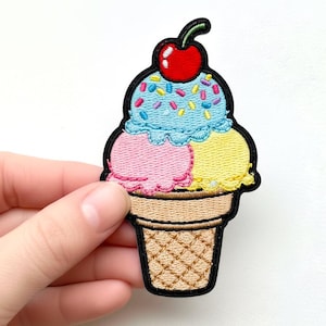 Könnte beinhalten: Gestickter Aufnäher mit einem Eiswaffel-Design. Das Eis hat drei Kugeln: rosa, gelb und blau, garniert mit Streuseln und einer roten Kirsche. Der Waffel-Kegel ist hellbraun und hat ein Waffelmuster.