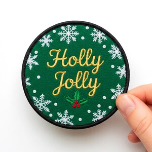 Puede incluir: Un parche redondo verde con las palabras "Holly Jolly" en letras doradas. Decorado con copos de nieve blancos y una ramita de acebo con bayas rojas. Borde negro.