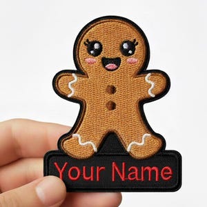 Peut inclure: Écusson en forme de bonhomme en pain d'épices marron avec un visage souriant, des détails de glaçage blanc et un bord noir. L'écusson a une base rectangulaire noire avec le texte "Your Name" en rouge. L'écusson est tenu par une main.