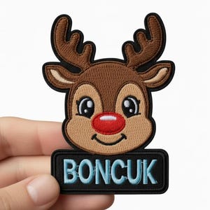 Puede incluir: Parche bordado con la cara de un reno de dibujos animados con una nariz roja, cuernos marrones y la palabra "BONCUK" en letras azules sobre un fondo negro. El parche está en la mano de una persona.