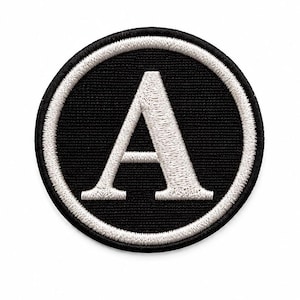 May include: A circular embroidered patch featuring a black background with a white border and a large white capital letter A centered in the middle. The design is stitched with a visible thread texture throughout the entire circular shape.