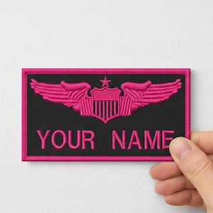 Könnte beinhalten: Ein schwarzer, gestickter Aufnäher mit rosa Rand und Design. Das Design zeigt Flügel, ein Schild und einen Stern. Der Text "YOUR NAME" ist rosa gestickt. Der Aufnäher ist rechteckig.