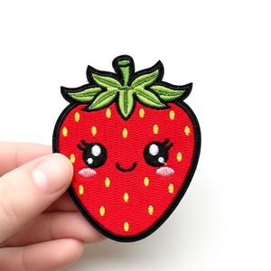 Peut inclure: Écusson de fraise rouge avec un visage souriant, des yeux noirs et des feuilles vertes. L'écusson a une bordure noire et des graines jaunes. L'écusson est tenu par une main.