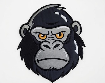 Könnte beinhalten: Ein Stoffaufnäher mit einem stilisierten Gorillakopf mit dunklem schwarzem Fell, einem hellgrauen Gesicht und leuchtend orangefarbenen Augen. Das Design enthält weiße Highlights auf der Stirn und an der Seite des Kopfes mit dicken schwarzen Stickrändern.