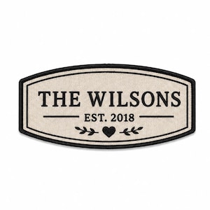 Puede incluir: Un parche rectangular con esquinas redondeadas, fondo de tela color crema y borde bordado en negro. El texto THE WILSONS está centrado sobre EST. 2018, seguido de un pequeño icono de corazón flanqueado por dos motivos de ramas con hojas.