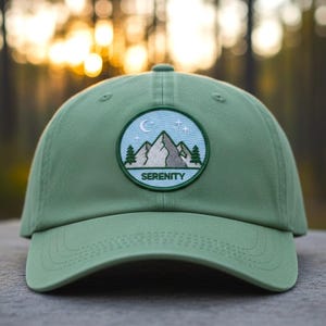 May include: A sage green baseball cap with a curved brim. The cap features a circular patch with a mountain scene, a crescent moon, and stars, with the word "SERENITY" below. The background is a blurred forest.