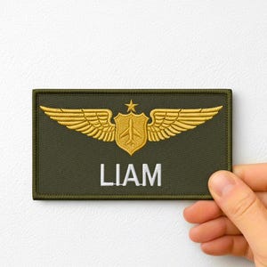 Könnte beinhalten: Olivgrüner Aufnäher mit goldgestickten Flügeln und einem Schilddesign. Der Name "LIAM" ist in Weiß gestickt. Der rechteckige Aufnäher hat einen gestickten Rand, ideal für Uniformen oder Kleidung.