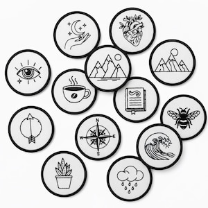 May include: A collection of round, white embroidered patches with black borders. Each patch features a different design, including a crescent moon and hand, a heart, mountains, an eye, a coffee cup, a book, an arrow, a compass, a bee, a wave, a plant, and rain.
