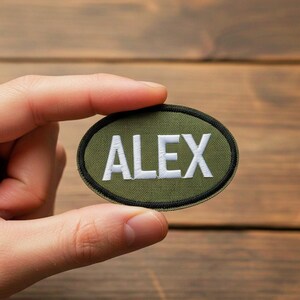Pode incluir: Um patch oval verde com o nome "ALEX" bordado em branco. O patch tem uma borda preta e é exibido em um fundo de madeira.