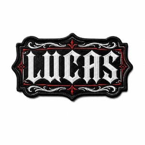 Pode incluir: Um emblema bordado retangular com fundo preto e borda decorativa. O nome LUCAS está bordado em letras brancas grandes de estilo gótico no centro, rodeado por detalhes intrincados de volutas e linhas em branco e vermelho.