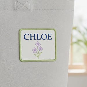 Peut inclure: Un sac fourre-tout gris clair avec un écusson brodé avec le nom "CHLOE" en bleu au-dessus d'un motif floral. L'écusson a une bordure vert clair et les fleurs sont violettes.