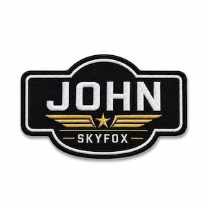 May include: A rectangular embroidered patch with rounded corners featuring a black background and a white border. The text JOHN is stitched in white, centered above a gold star flanked by gold wings, with the text SKYFOX below in white.