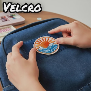 May include: A circular embroidered patch featuring an orange sun with radiating rays above stylised blue ocean waves. The patch is positioned on the front of a dark blue denim backpack, which has a rectangular pocket with a visible Velcro strip.