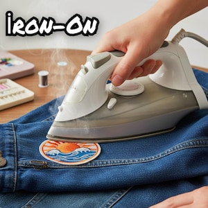 Pu&ograve; includere: Un ferro da stiro a vapore bianco e grigio preme una toppa circolare ricamata su un tessuto denim blu. La toppa presenta un grafico di un sole arancione che sorge su onde oceaniche blu e bianche. Il testo IRON-ON &egrave; in lettere nere.