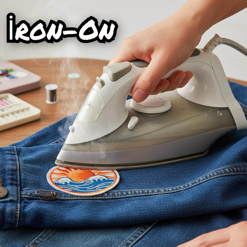 Pu&ograve; includere: Un ferro da stiro a vapore bianco e grigio preme una toppa circolare ricamata su un tessuto denim blu. La toppa presenta un grafico di un sole arancione che sorge su onde oceaniche blu e bianche. Il testo IRON-ON &egrave; in lettere nere.