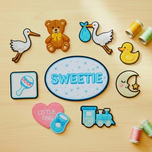 Może przedstawiać: Kolekcja haftowanych naszywek o tematyce dziecięcej, w tym miś, bocian, kaczka, księżyc, grzechotka, serce, pociąg i owalna naszywka z napisem "SWEETIE". Naszywki są w różnych kolorach.